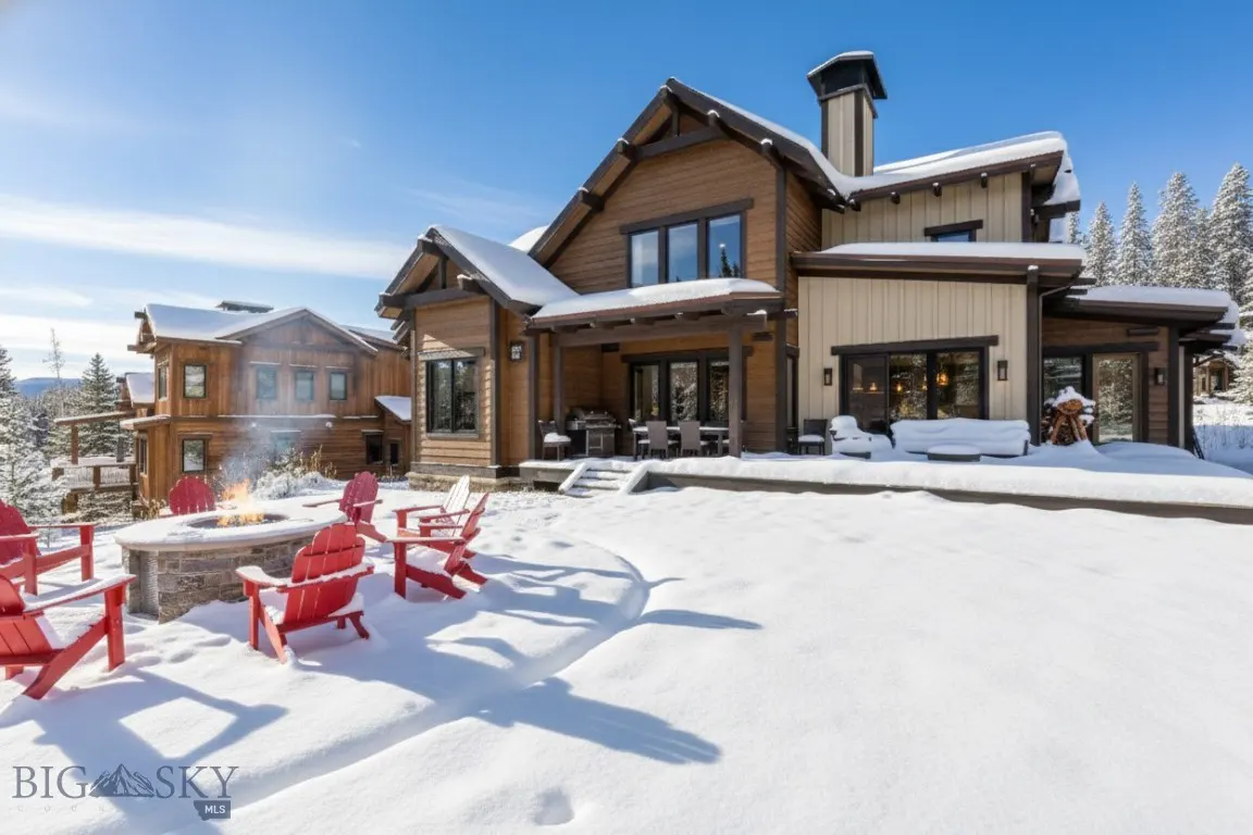 A snow-covered luxury home with a stone fire pit and red Adirondack chairs. Pine trees and a blue sky are in the background.