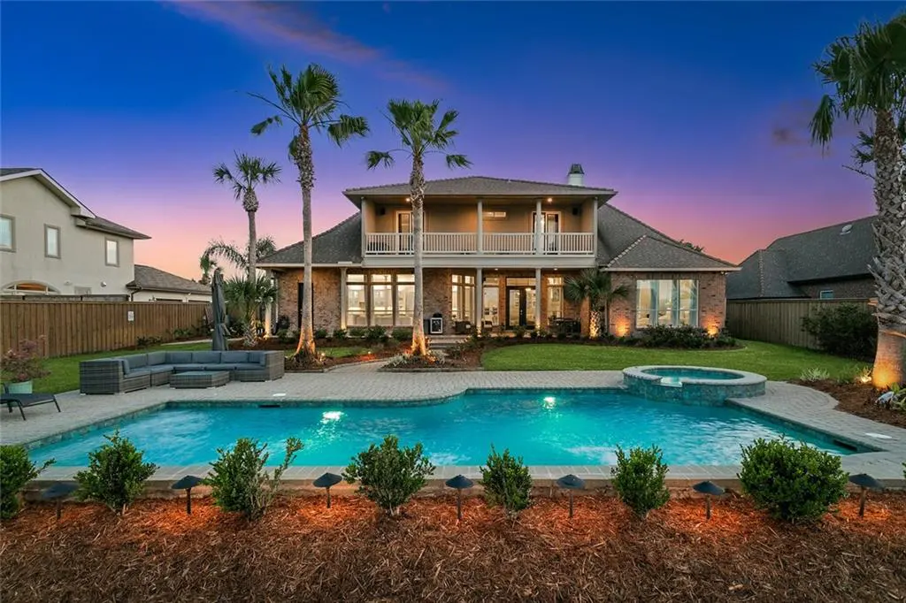 Two-story brick house with a pool, hot tub, and palm trees at dusk. A gray outdoor sofa sits near the pool.
