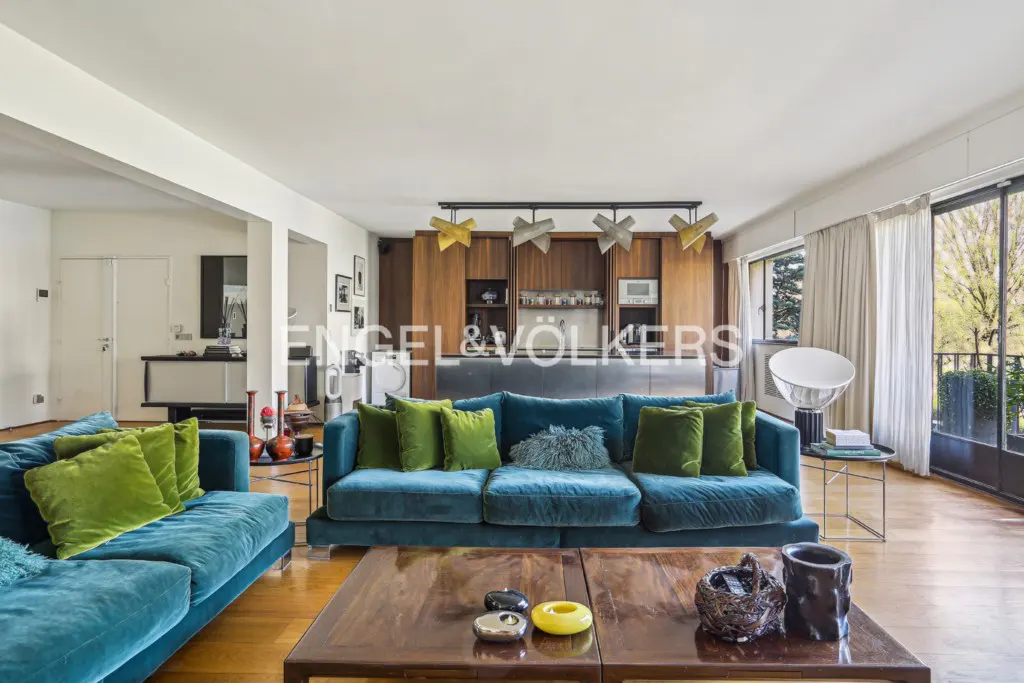 Living room with blue velvet sofas, green pillows, wood floors, and a dark wood coffee table. Kitchen in the background.
