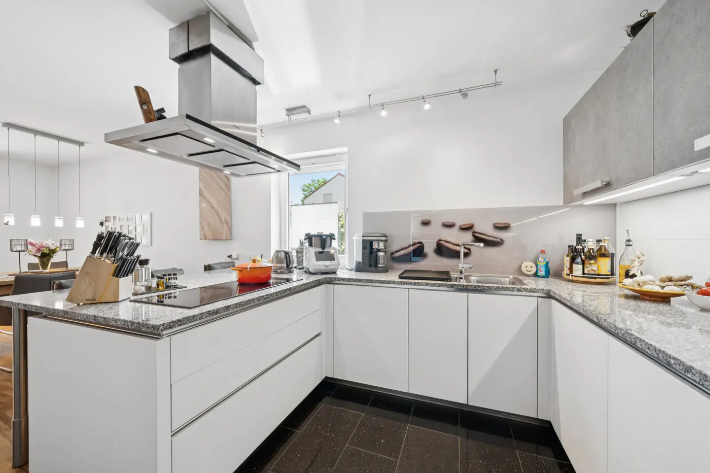 Bright kitchen with white cabinets, granite countertops, and black tile floor. Stainless steel range hood above island. Dining area visible.