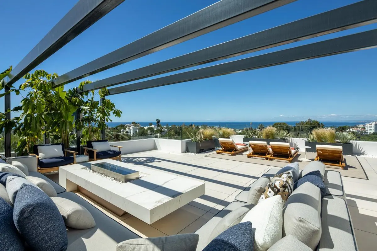 Modern rooftop terrace with a white fire pit, lounge seating, and a pergola overlooking the ocean under a clear blue sky.