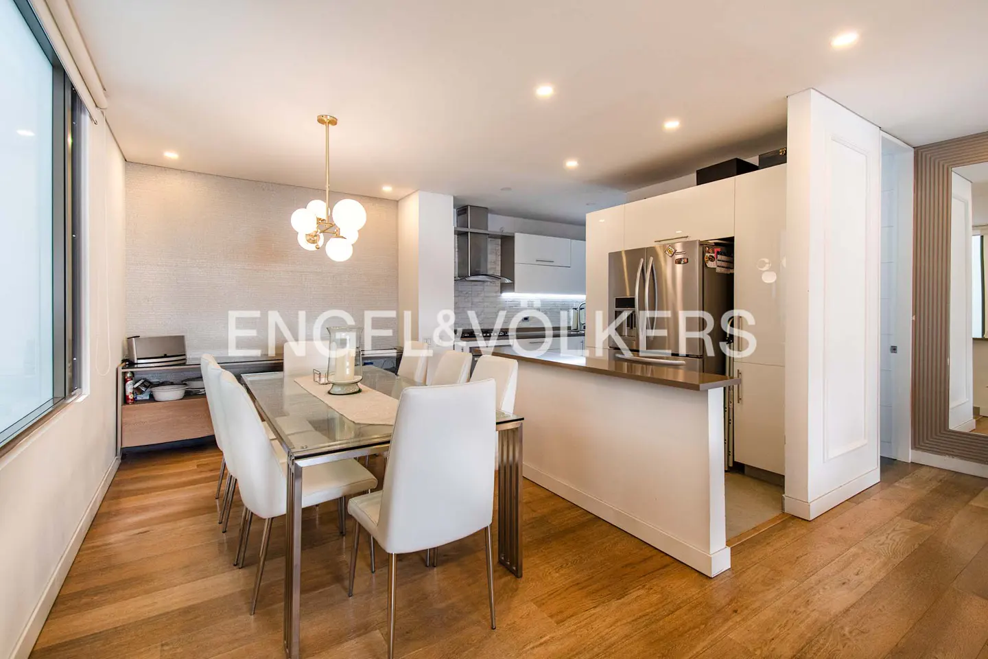 Open-concept dining and kitchen area with a glass table, white chairs, and stainless steel appliances. Hardwood floors and modern lighting.