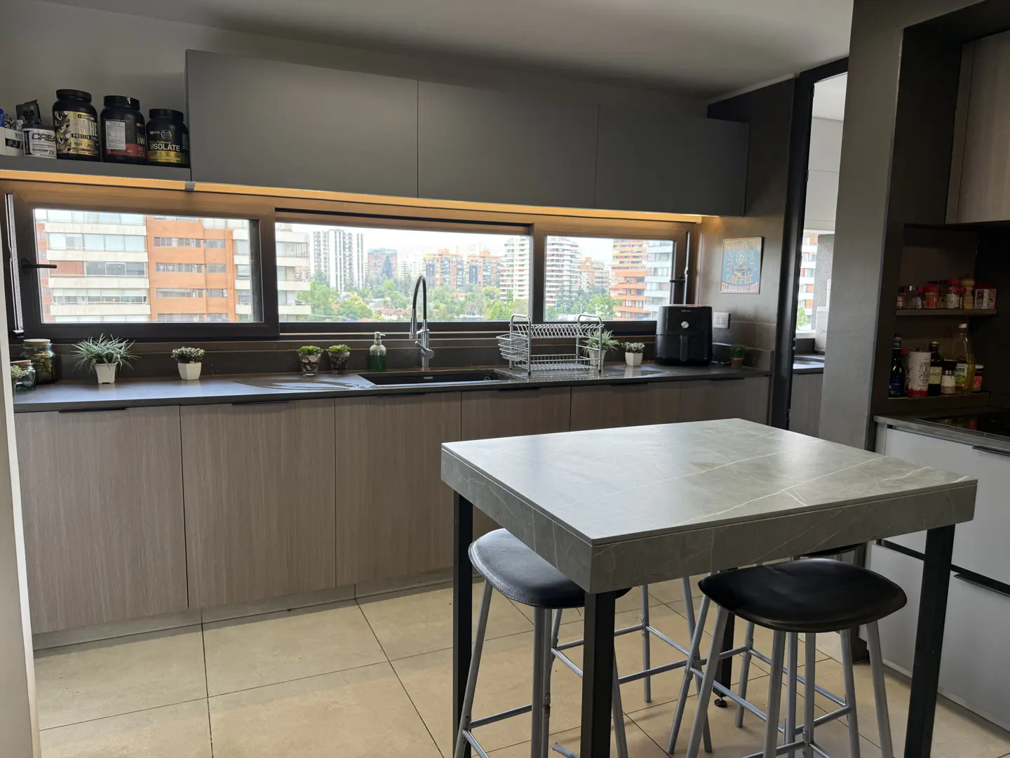 Modern kitchen with light wood cabinets, grey stone island, two black stools, and a long window overlooking city buildings.