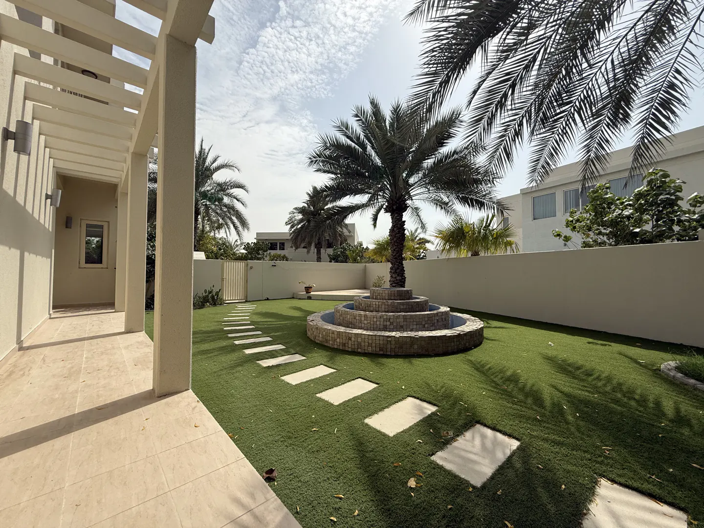 A backyard with a palm tree in the center, surrounded by a tiered stone fountain. A stone path leads to a gate in the white wall.