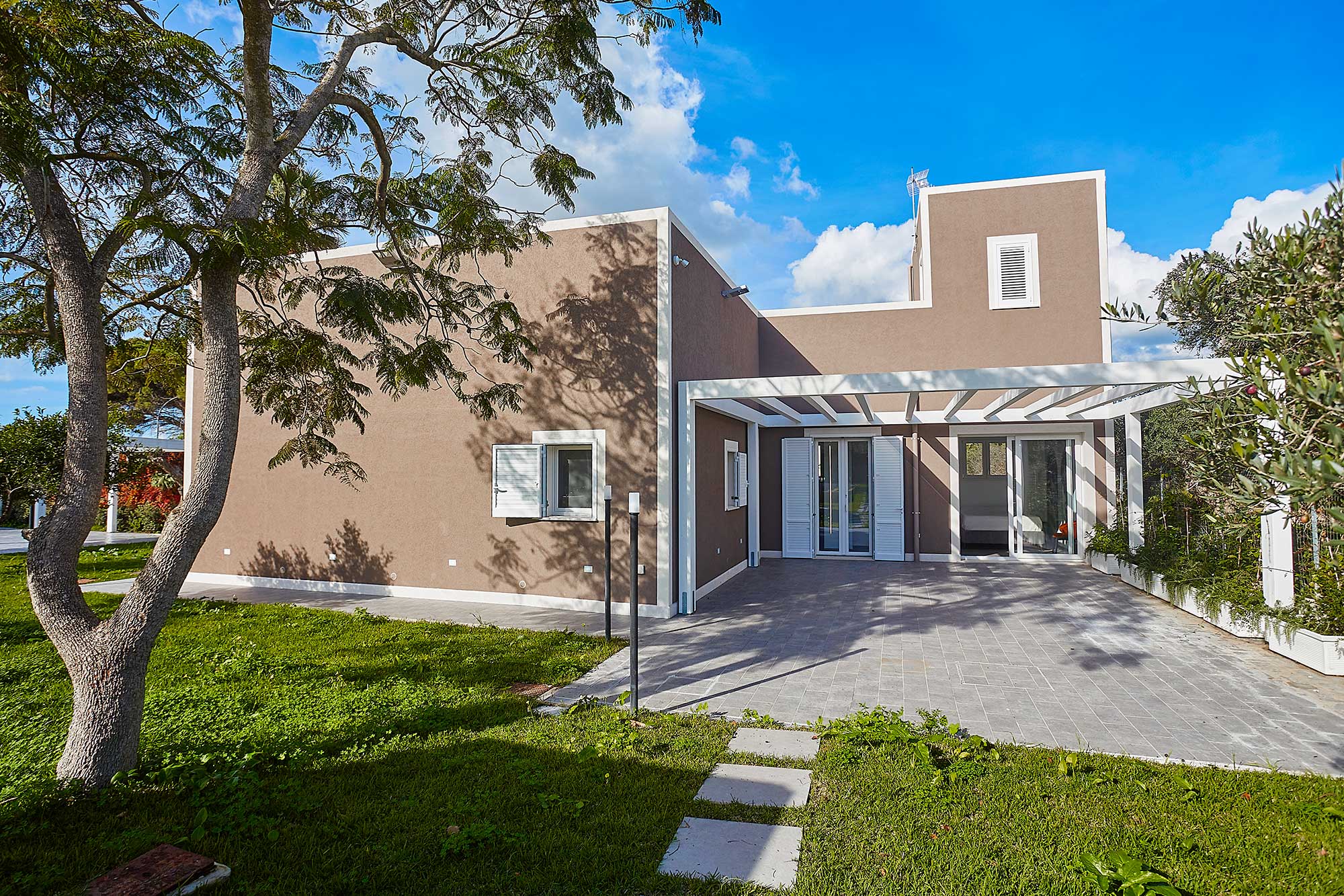 Exterior view of a modern, brown house with a white pergola, stone patio, and green lawn under a blue sky.