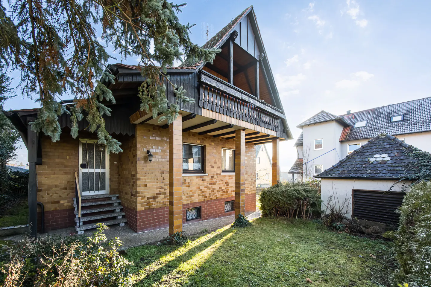 Two-story brick house with a balcony and a peaked roof, surrounded by a green lawn and trees.