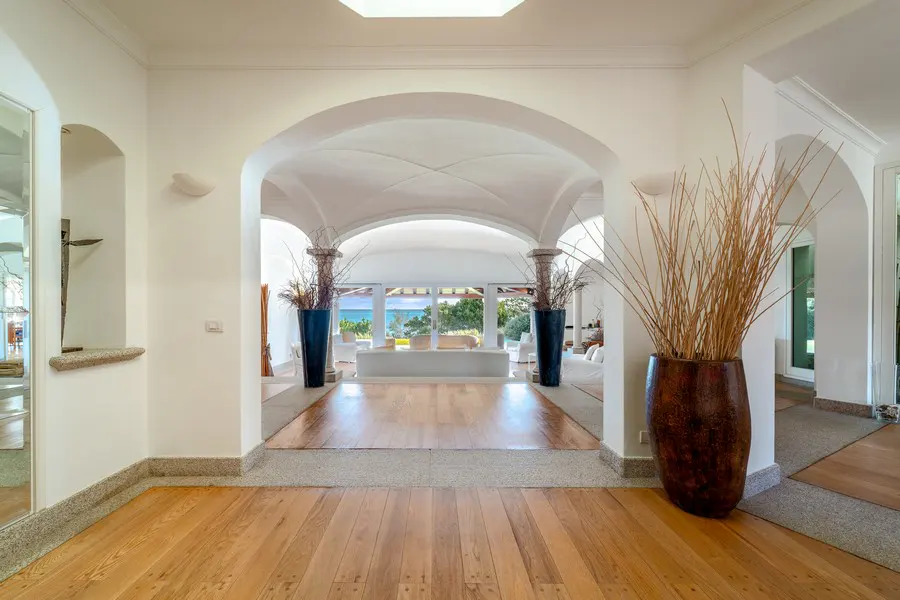 Elegant foyer with wood floors, white walls, and arched doorways leading to a bright living area with ocean views. Vases with tall branches add a natural touch.