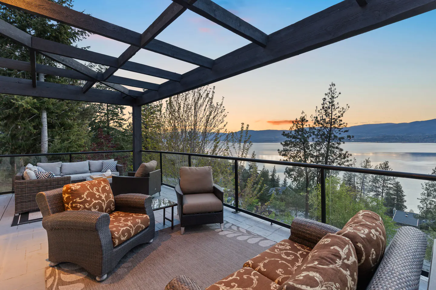 Outdoor patio with wicker furniture, glass railing, and a pergola. A lake and sunset are visible in the background.