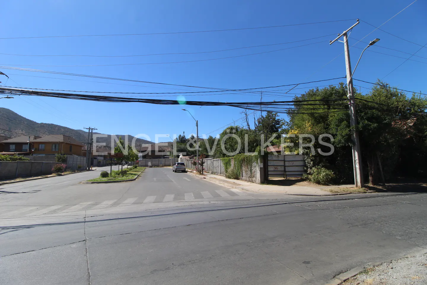Street view of a residential area with a car on the road, houses, trees, and a clear blue sky.