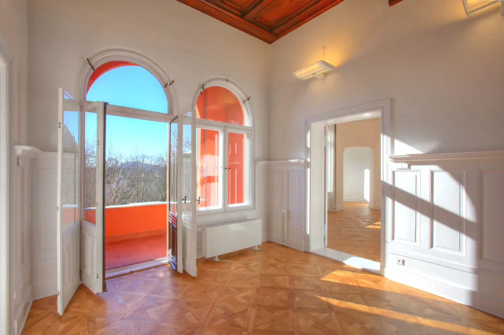Bright, empty room with parquet floors, white walls, and orange trim. Open doors lead to a balcony with a view of trees.