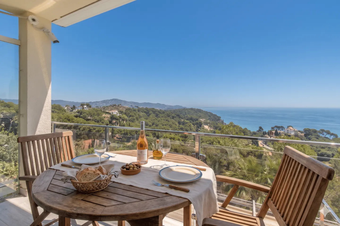 Outdoor dining on a balcony with a sea view. A wooden table is set with wine, bread, olives, and plates.