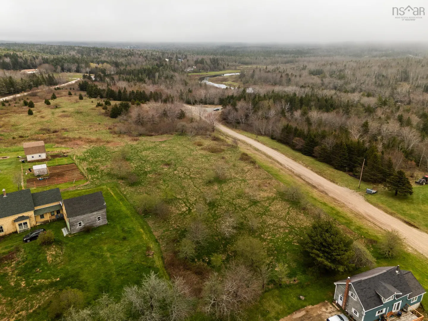 Aerial view of a rural property with a dirt road, fields, and houses under a cloudy sky. Forest in the background.