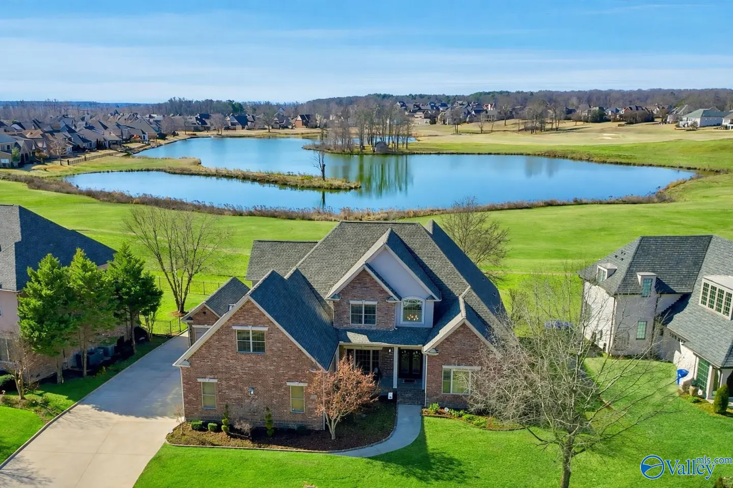 Aerial view of a two-story brick house with a gray roof, green lawn, and a lake in the background on a sunny day.