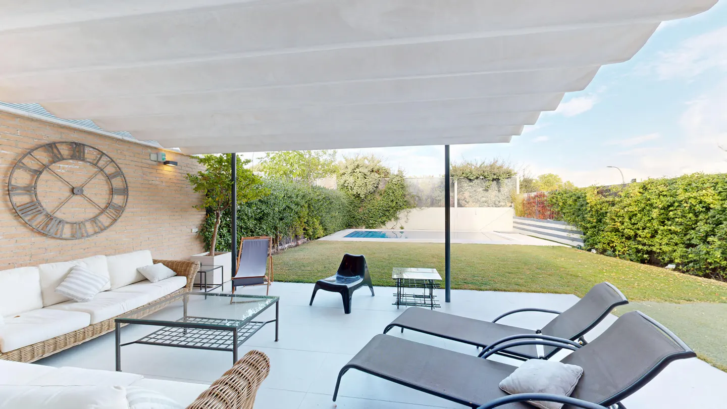 Outdoor patio with lounge chairs, sofa, and a large clock on a brick wall. A green lawn and pool are visible in the background.