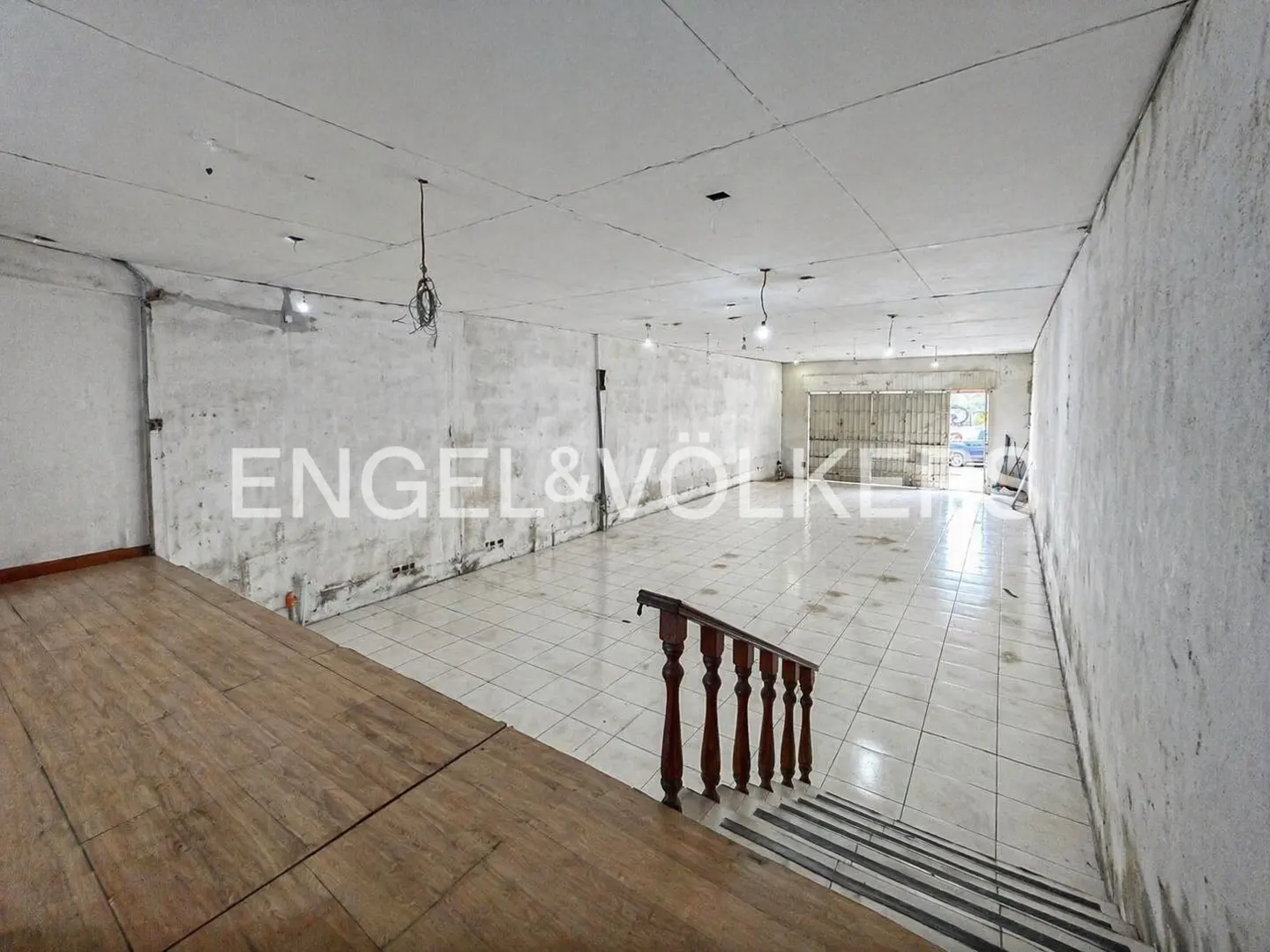 Unfinished commercial space with white tile floor, concrete walls, and wooden stairs with a dark brown railing. Engel & Völkers logo is visible.