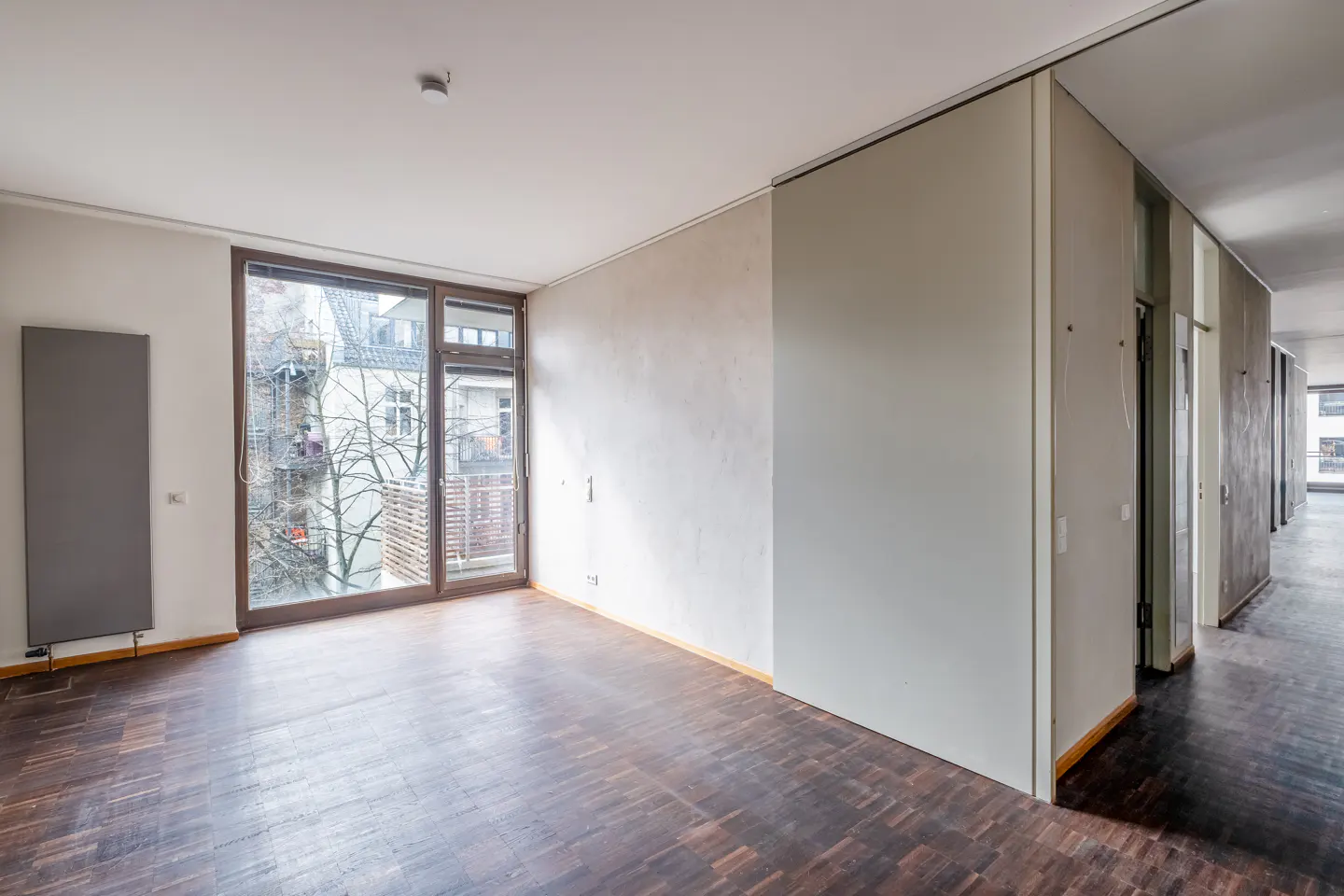 An empty room with dark wood floors, white walls, and a large window. Outside the window is a view of trees and buildings.
