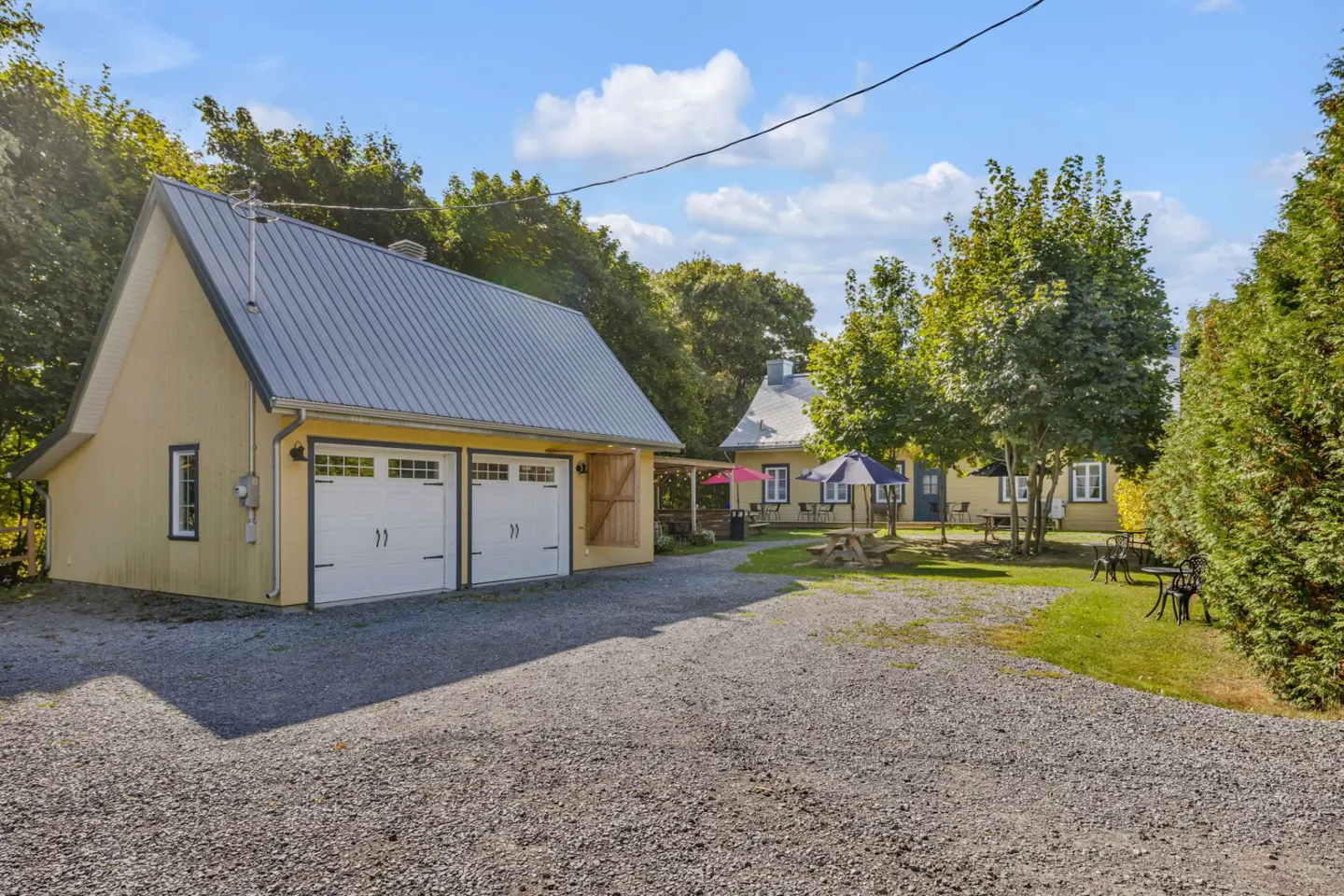 Exterior view of a yellow building with a gray roof and two white garage doors, surrounded by trees and a gravel driveway.