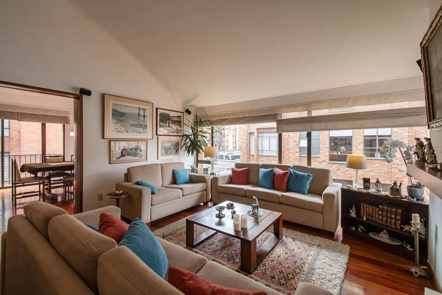 Living room with beige sofas, blue and red pillows, wood floors, and a view of a brick building.