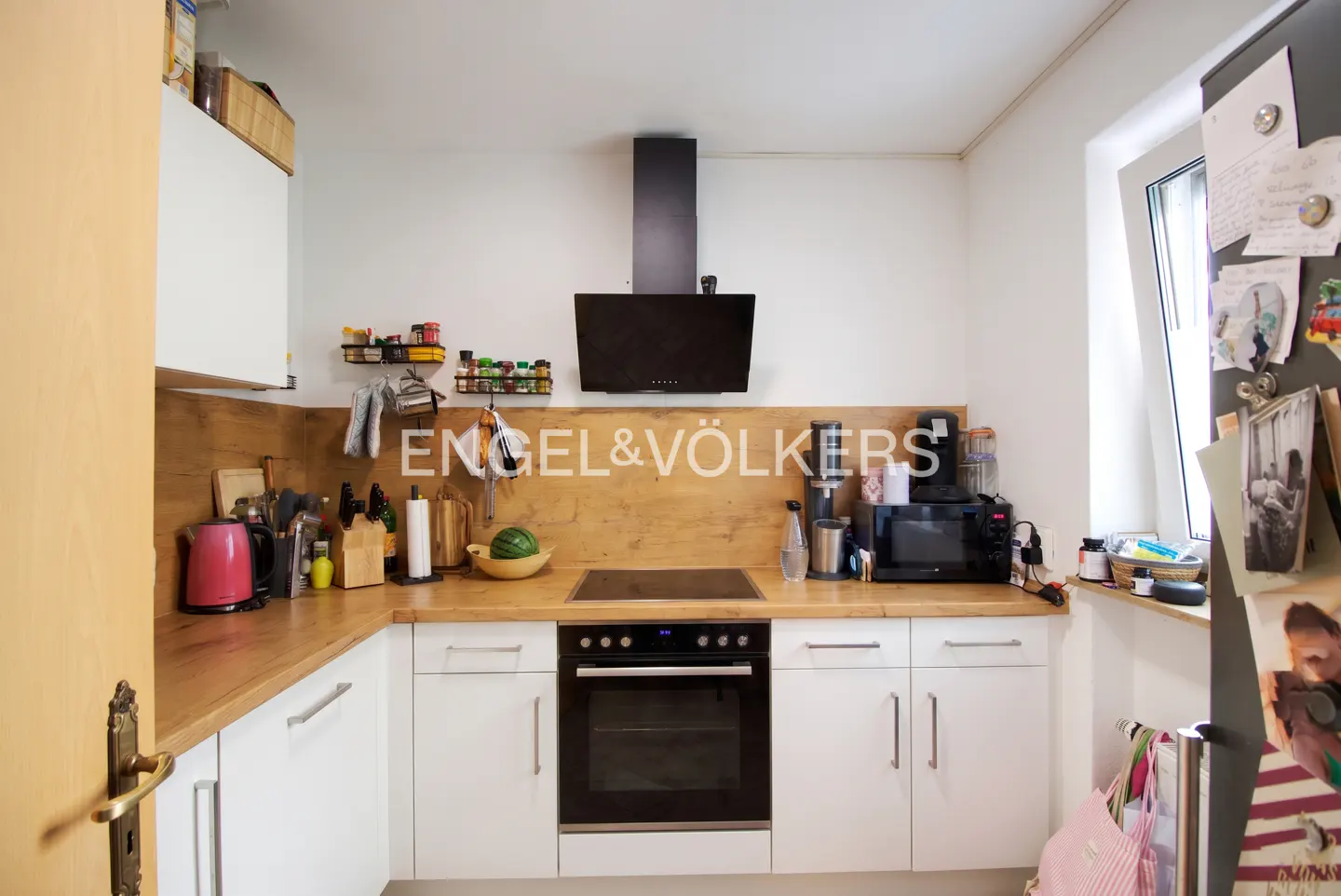 A bright kitchen with white cabinets, a wood countertop, and a black range hood. The Engel & Völkers logo is on the backsplash.