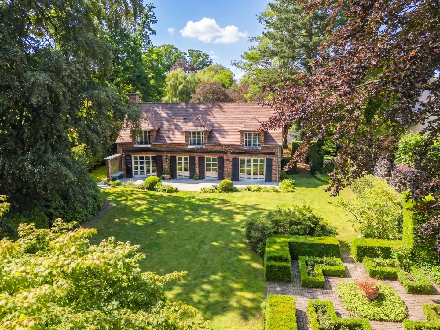 A two-story brick house with a tiled roof, large windows, and a spacious green lawn surrounded by lush trees and hedges.
