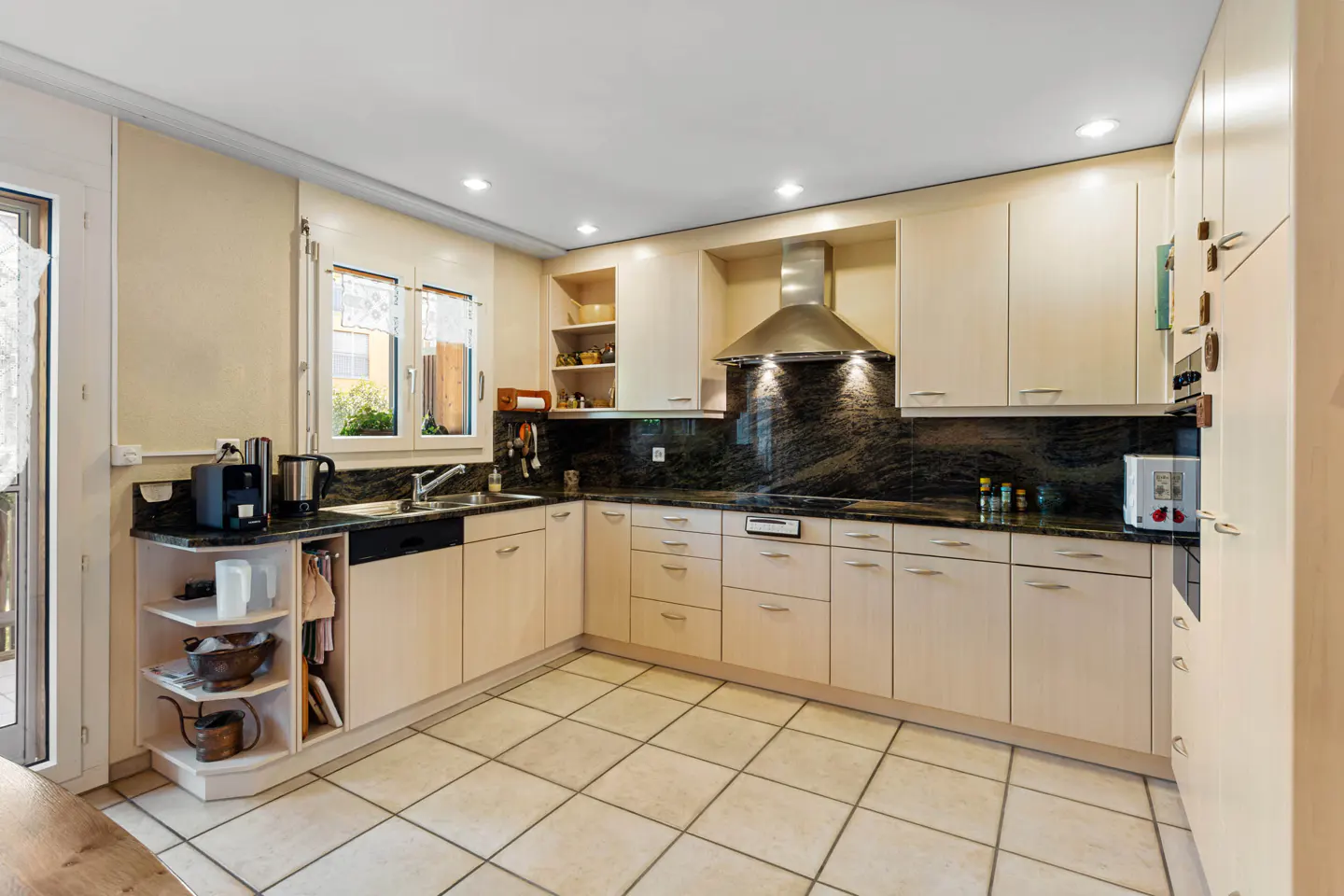 Bright kitchen with beige cabinets, black granite countertops, and stainless steel range hood. Cream-colored tile floor and open shelving.