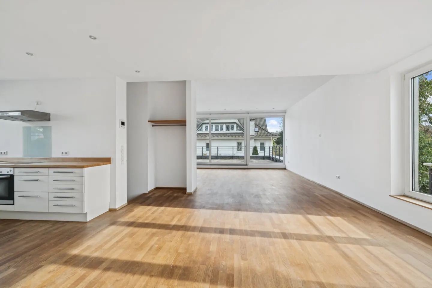 Bright, empty apartment with wood floors, white walls, and a kitchen area. A large window offers a view of a house and greenery.