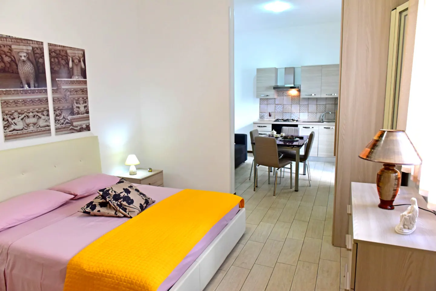 A bright studio apartment with a bed, kitchen, and dining area. The bed has pink sheets and a yellow blanket. A lamp sits on a nightstand.