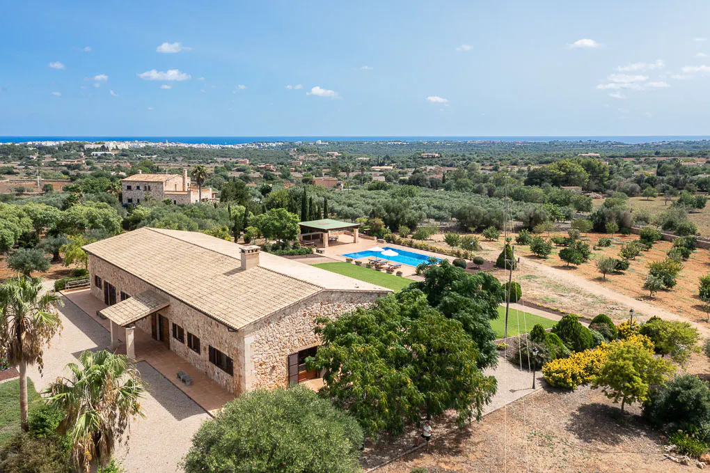Aerial view of a stone house with a tile roof, a blue pool, green trees, and the ocean in the background.
