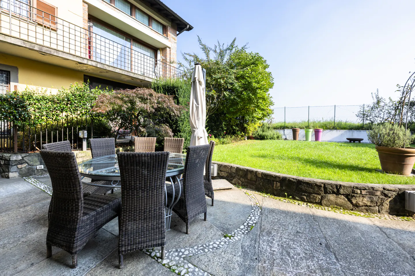 Outdoor patio with a glass table and wicker chairs, a green lawn, and a yellow building in the background.