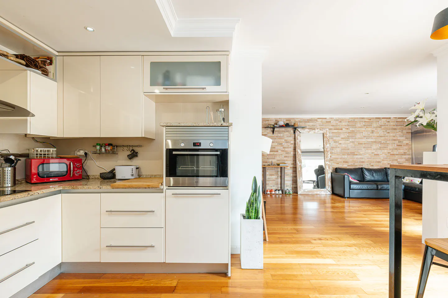 Open-concept kitchen and living room with wood floors. White cabinets, stainless steel oven, and red microwave in the kitchen. Black sofa in living room.