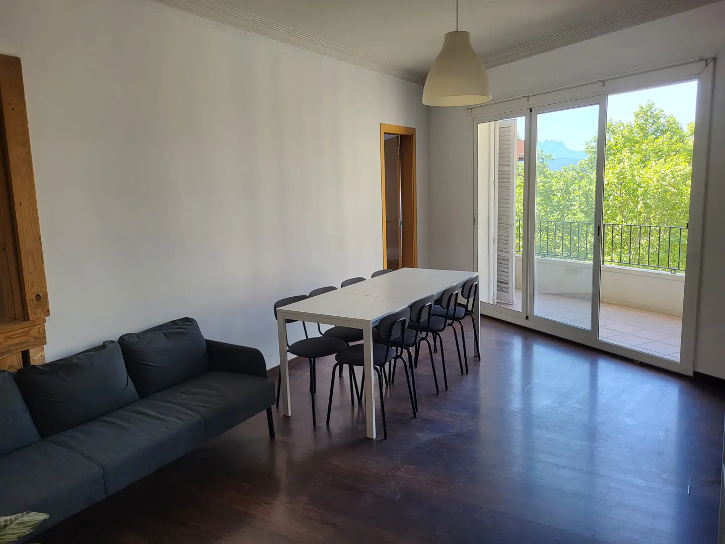 Bright room with a dark wood floor, a gray sofa, a white table with black chairs, and a balcony with a view of green trees.