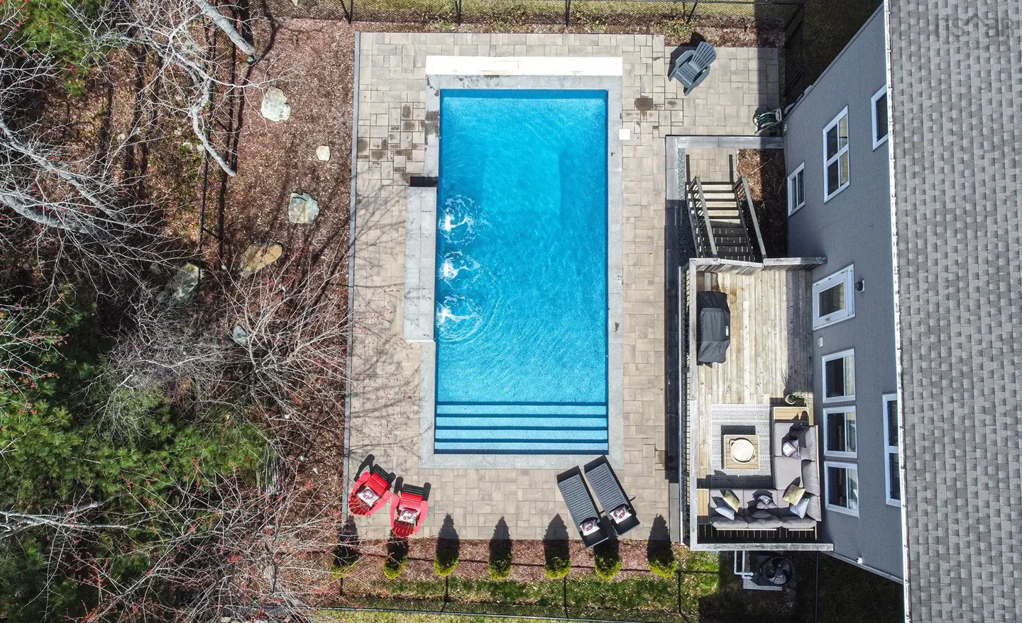 Aerial view of a backyard with a blue rectangular pool, patio furniture, a deck, and a gray house.