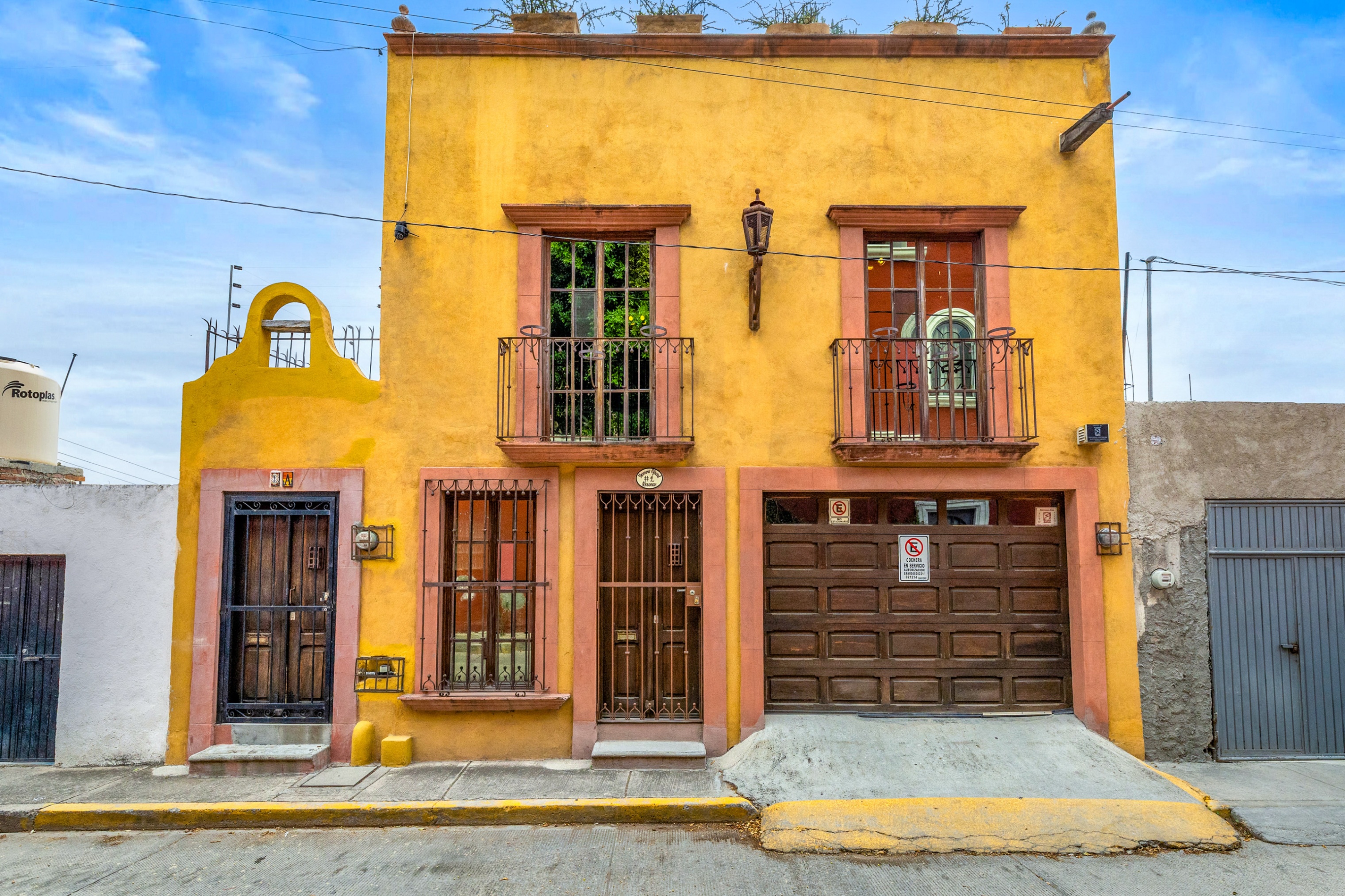 A two-story yellow house with pink trim, a brown garage door, and wrought iron balconies under a blue sky.