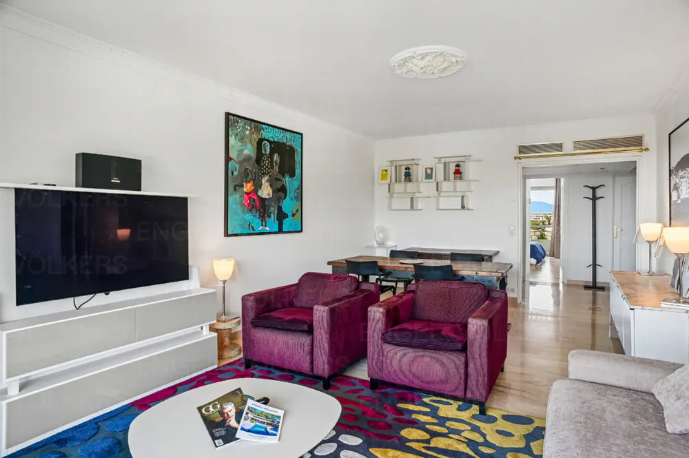 Bright living room with purple armchairs, a colorful rug, and a white coffee table with magazines. A TV and dining table are visible.