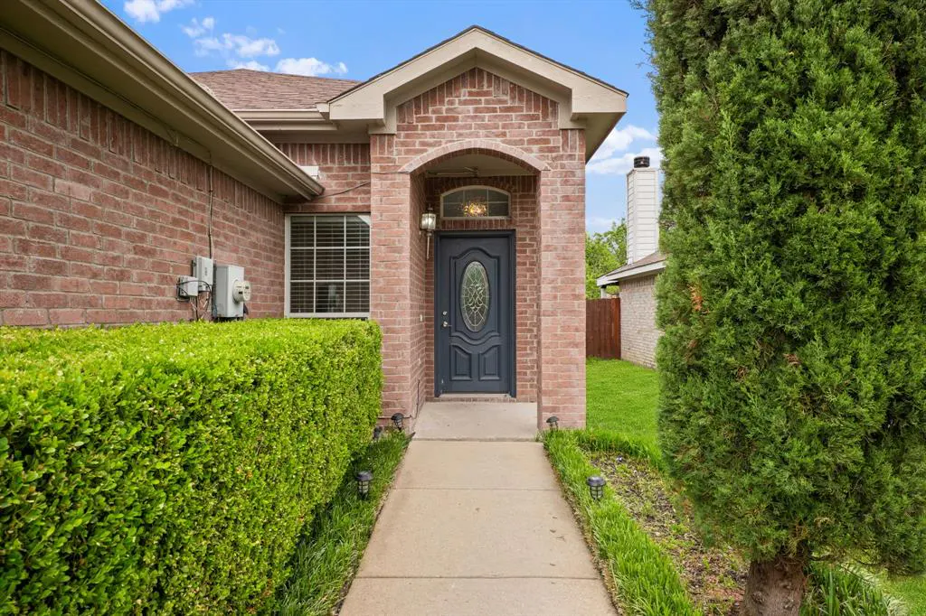 Brick house exterior with a gray front door, oval window, and arched entryway. A concrete walkway leads to the entrance, flanked by green bushes.