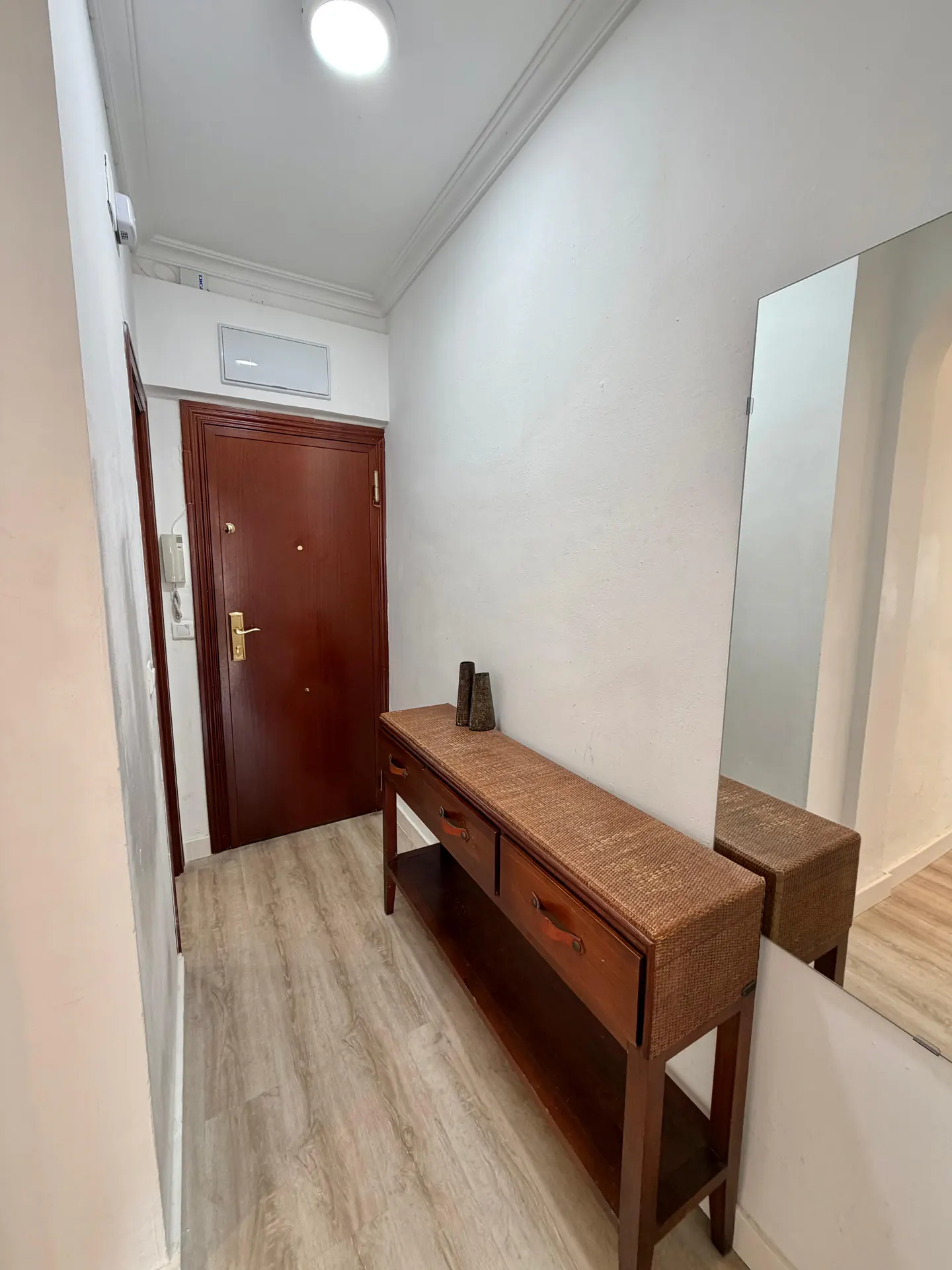 Entryway with a brown door, light wood floors, and a console table with drawers and a shelf. A mirror reflects the table.