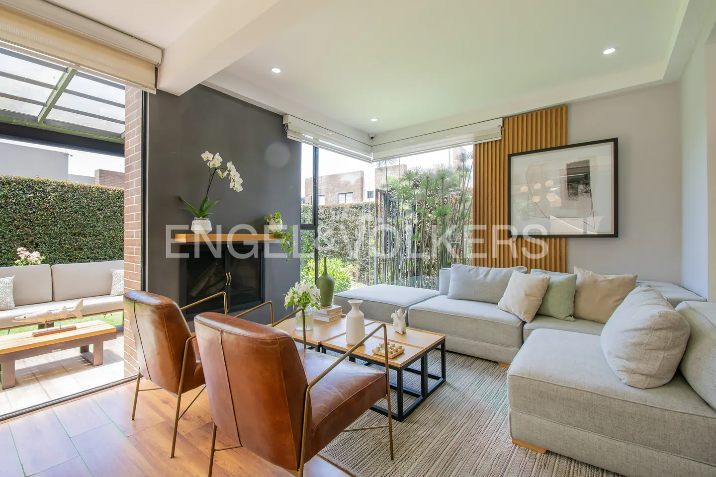 A bright living room with a gray sectional sofa, two brown leather chairs, and a view of a green garden.