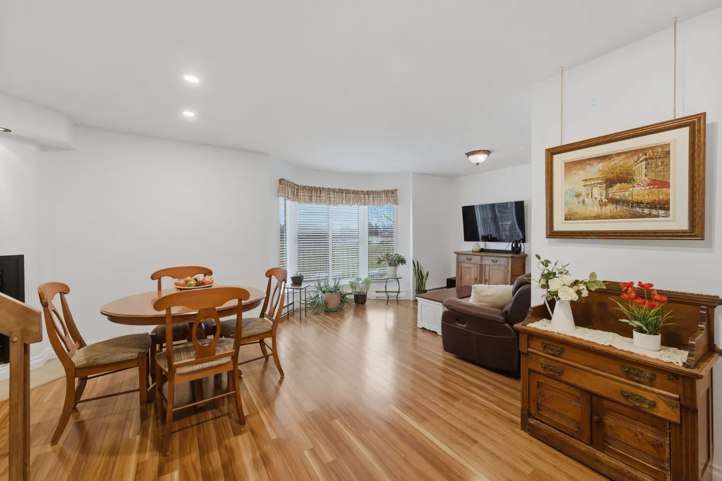 Bright living room with wood floors, a dining table with four chairs, a bay window, and a brown leather recliner. A framed painting hangs on the wall.