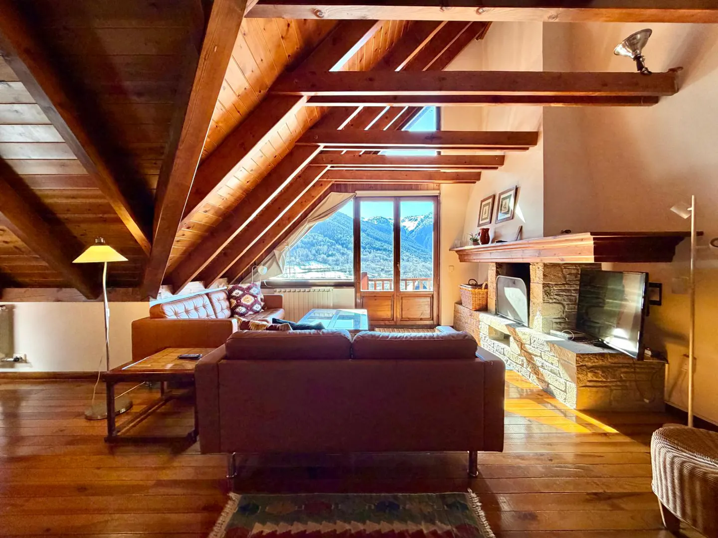 Attic living room with wood floors, exposed beams, and a stone fireplace. A balcony offers a view of snow-capped mountains.
