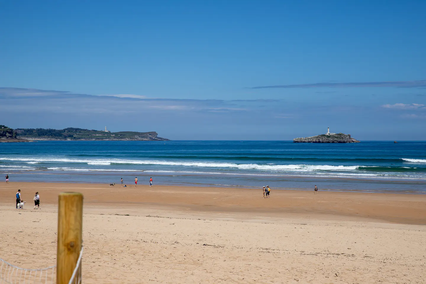 Sandy beach scene with blue sky, ocean, and distant islands. People stroll along the shore, enjoying the sunny day.