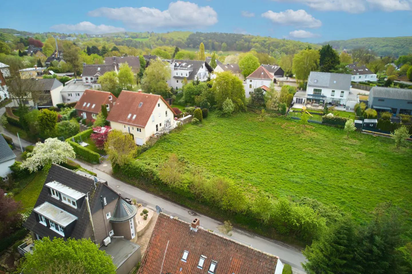 Aerial view of a residential area with houses, trees, and a large green field under a blue sky with clouds.