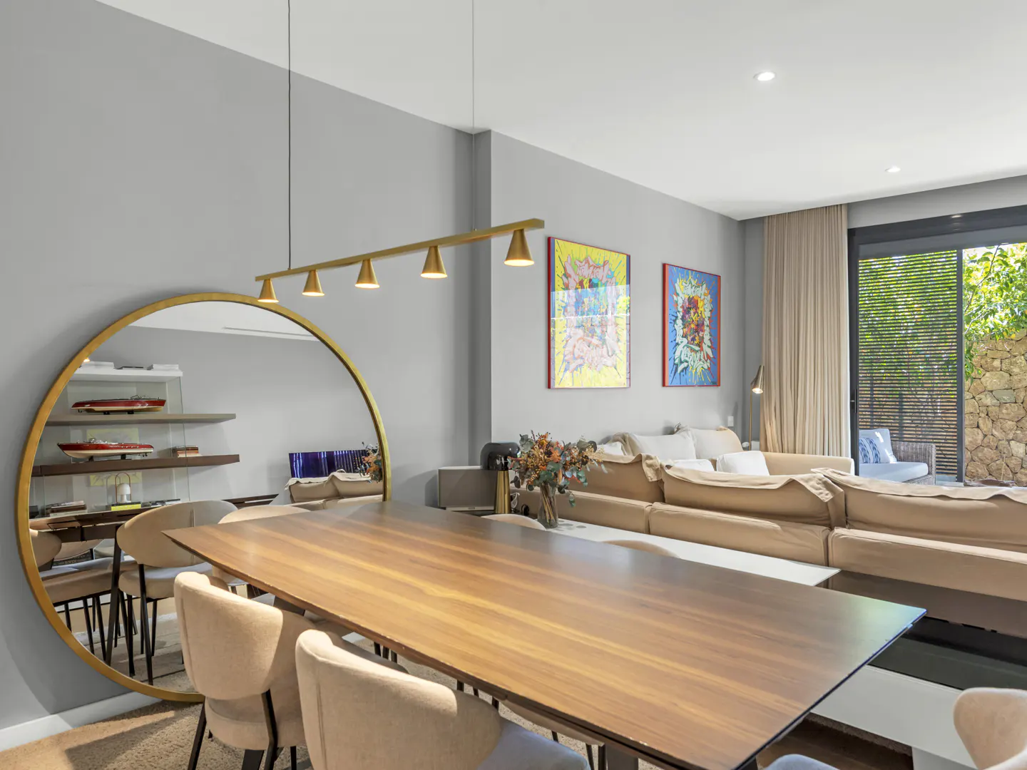 Open-concept living and dining area with a long wood table, beige chairs, and a large round mirror. Artwork and a tan sofa are in the background.
