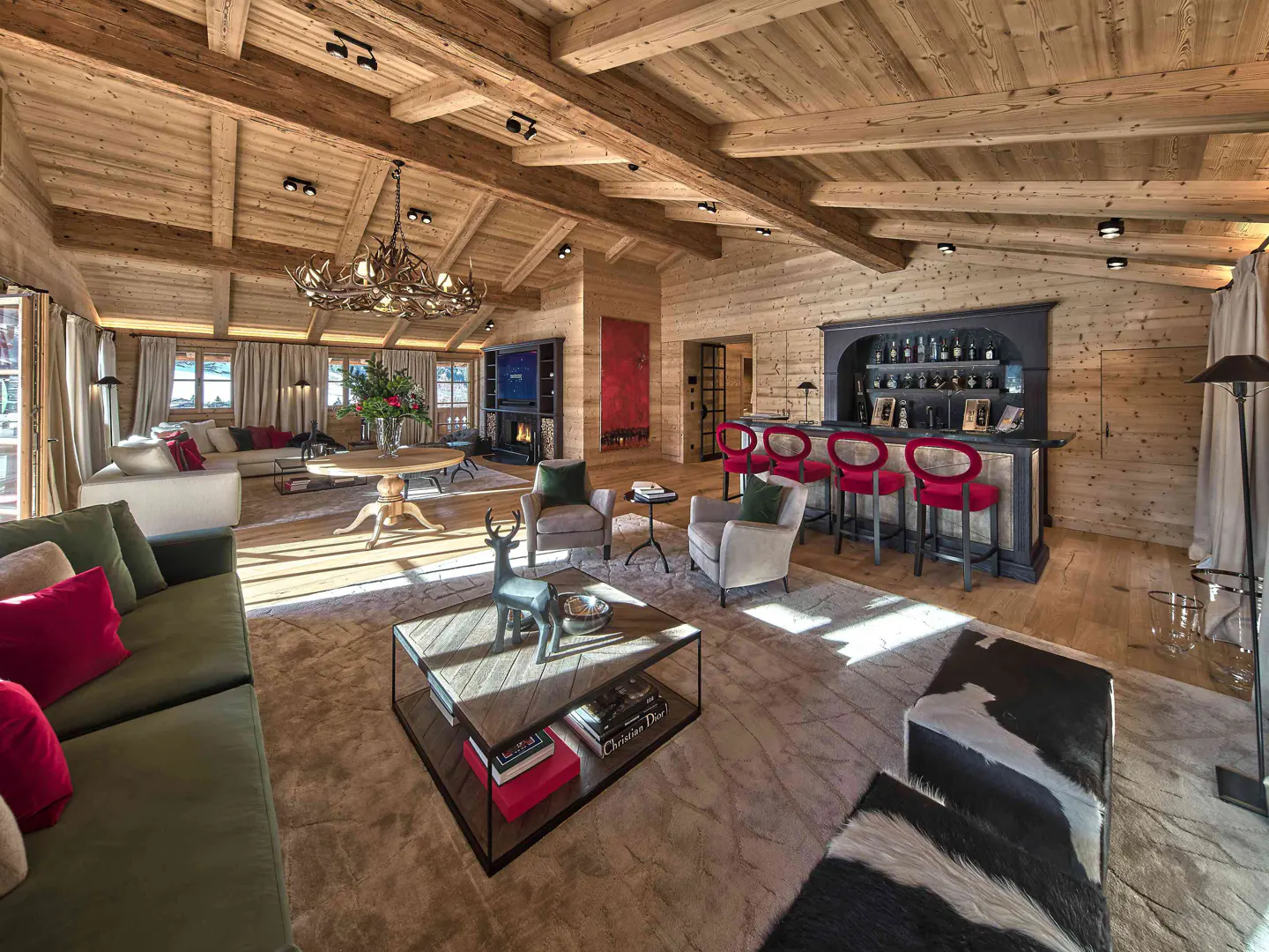 Luxury rustic living room with wood-paneled walls, antler chandelier, green sofas, and a bar with red stools.