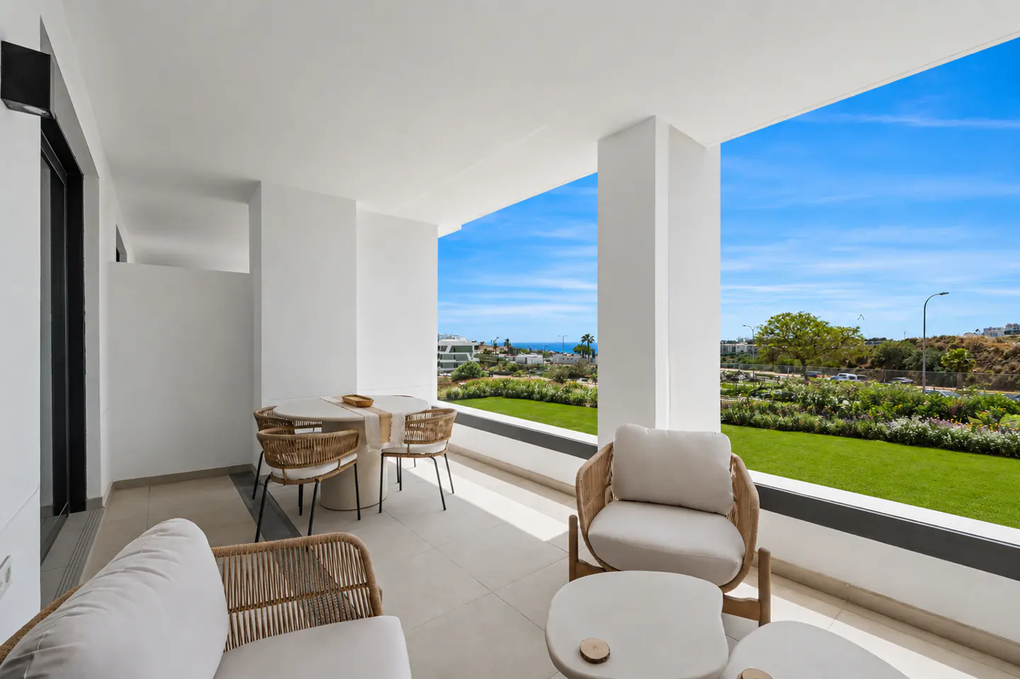 A bright, modern balcony with wicker furniture, a table set for dining, and a view of the ocean and green landscape.