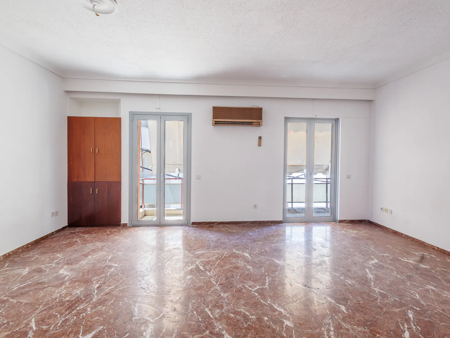Empty room with red marble floor, white walls, two glass doors to balconies, and a brown cabinet.