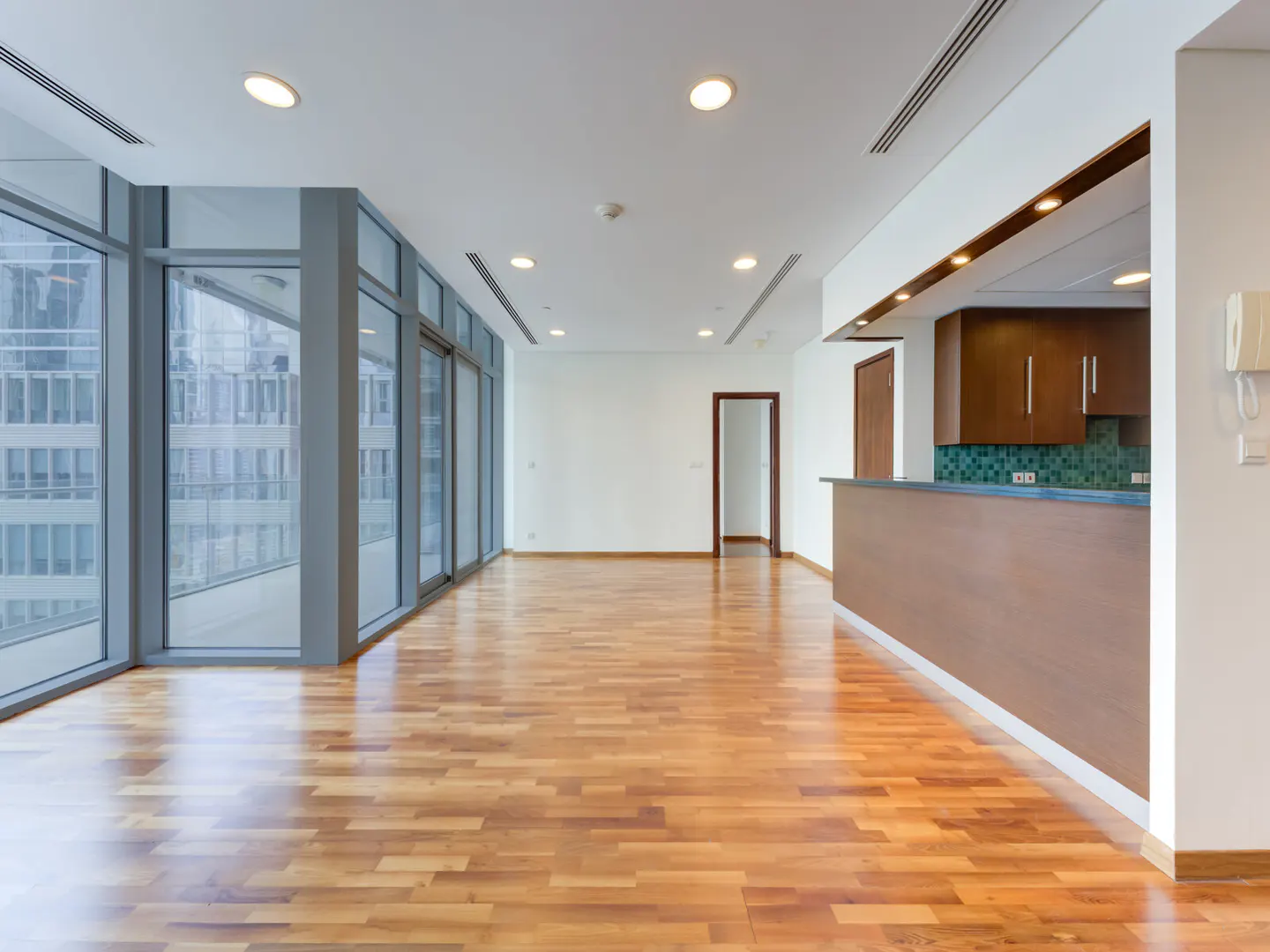 An empty, modern apartment with wood floors and floor-to-ceiling windows. A kitchen area with dark wood cabinets is visible.