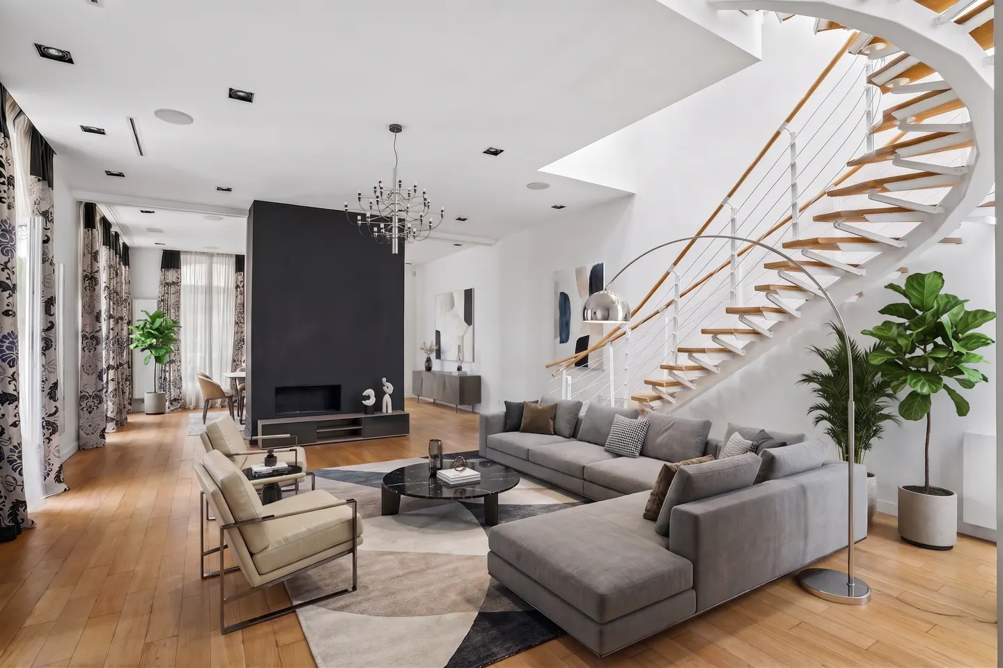 Bright, modern living room with a gray sectional sofa, round table, and spiral staircase. Hardwood floors and a black accent wall add contrast.
