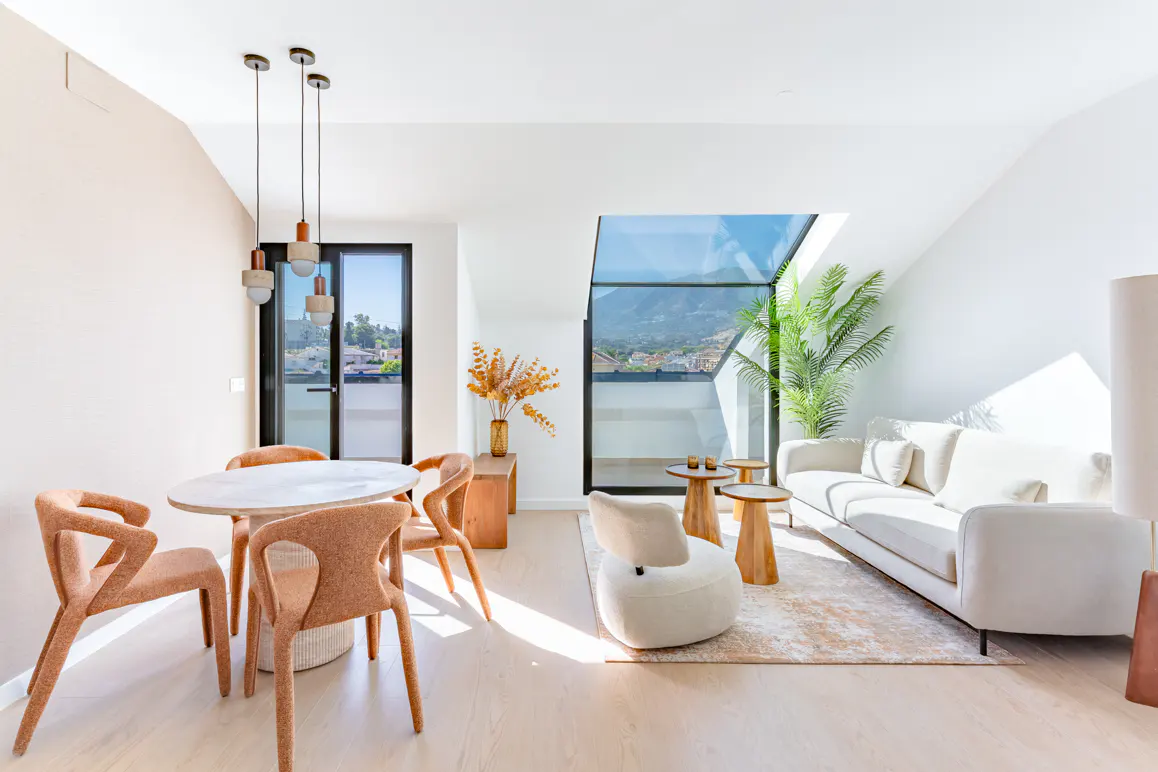 Bright, modern open-plan living area with a dining set, white sofa, and large windows overlooking a mountain landscape.
