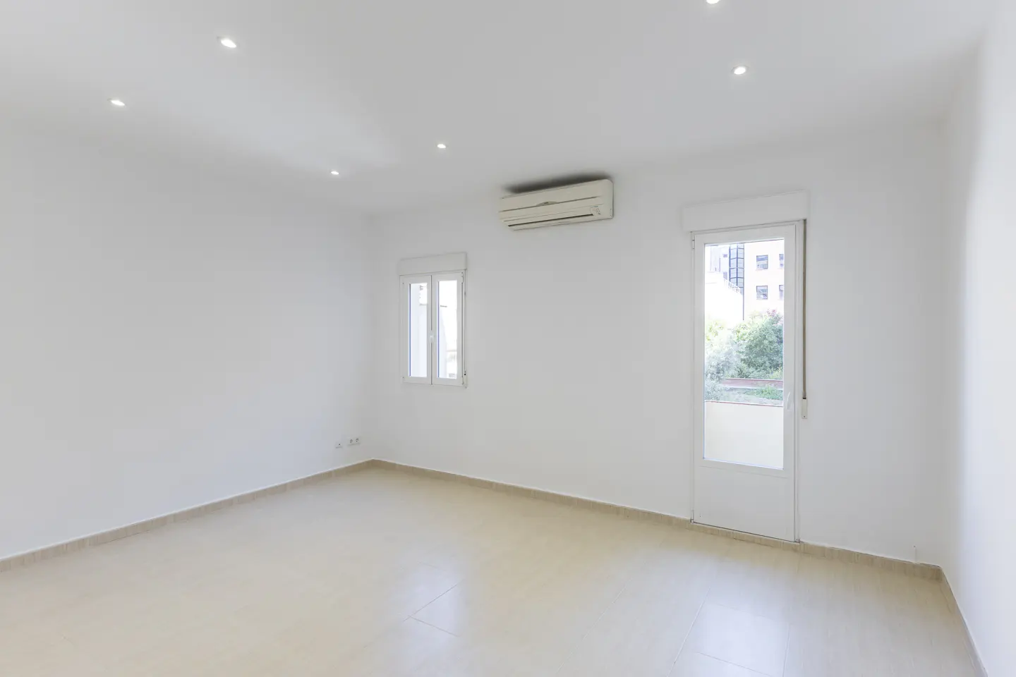Bright, empty room with white walls, beige tile floor, window, door to balcony, and air conditioner.
