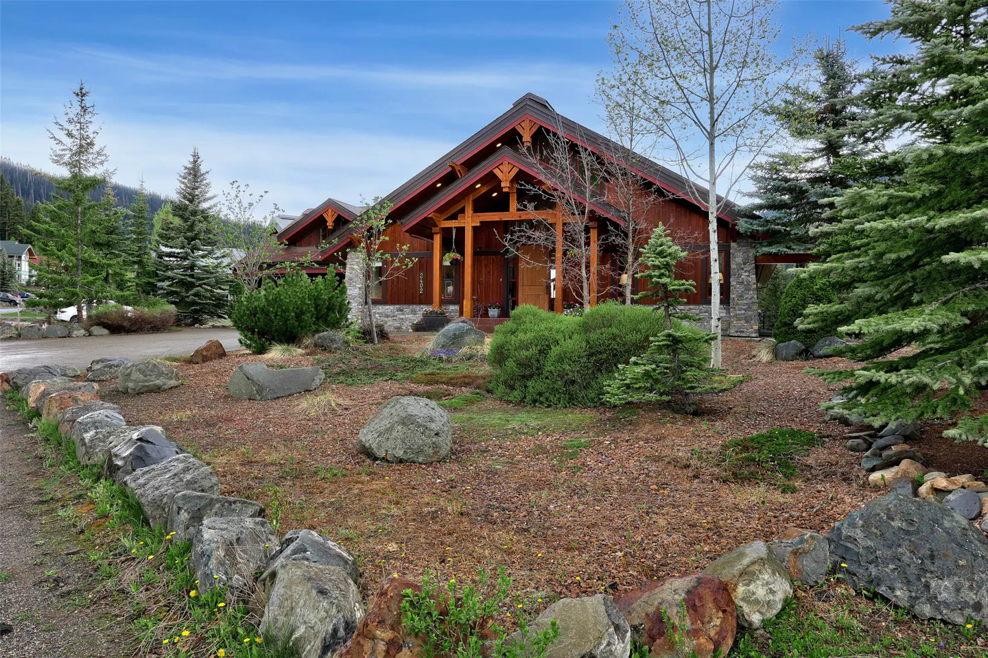 A brown, wood-sided house with a dark roof and stone accents, surrounded by trees and a rock garden.